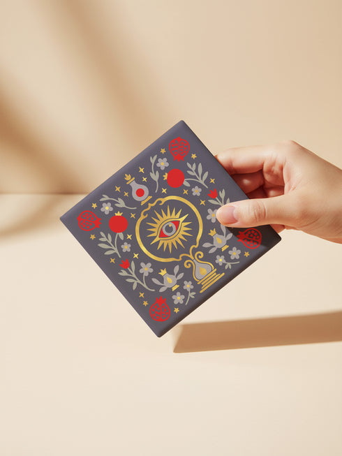 A square ceramic drink coaster with a smooth, matte charcoal grey finish. The surface features a symmetrical, intricate design in metallic gold, deep garnet red, and light grey. In the center is a golden All-Seeing Eye with a red iris, radiating light. The eye is framed by a golden circle and flanked by botanical elements, including red pomegranates and grey flowers with gold details. The design has a folk art, gothic botanical style.